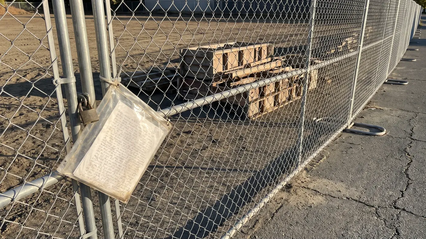 A padlocked chain link construction fence around a cleared jobsite in low late afternoon sun, with a weathered document pouch zip tied to the gate panel, stacked concrete forms and pallets on the dirt behind it, and long fence shadows across cracked asphalt.