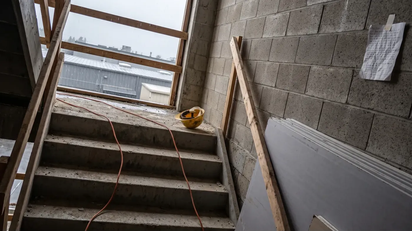 An unfinished concrete stairwell in a commercial building under construction on a gray rainy morning, with a dusty hard hat on the top step, an orange cord running down the treads, drywall stacked against the block wall, and a handwritten sheet taped up at the landing.